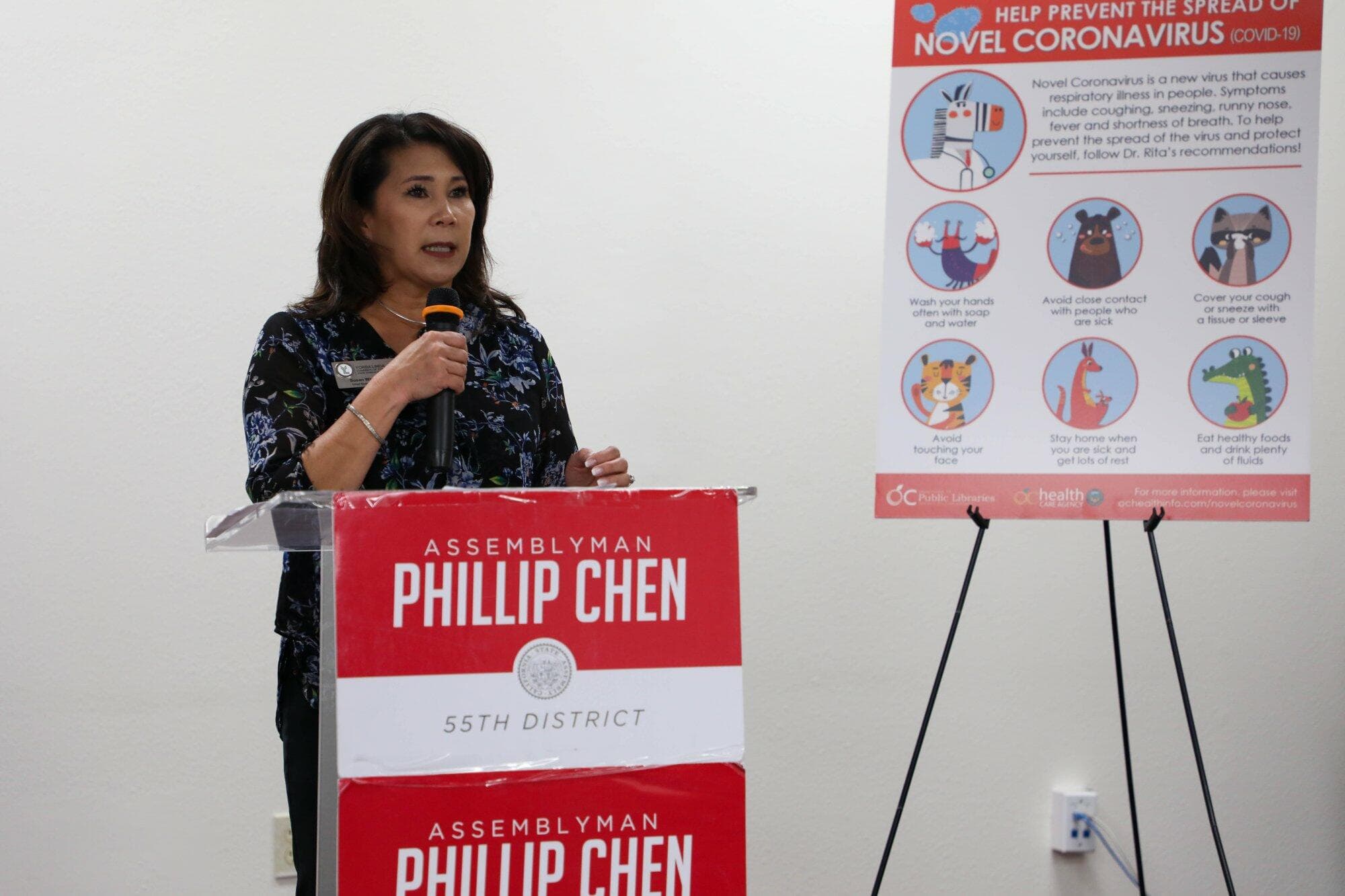 Yorba Linda Chamber of Commerce CEO Susan Wan-Ross speaking at the podium beside a COVID-19 prevention poster