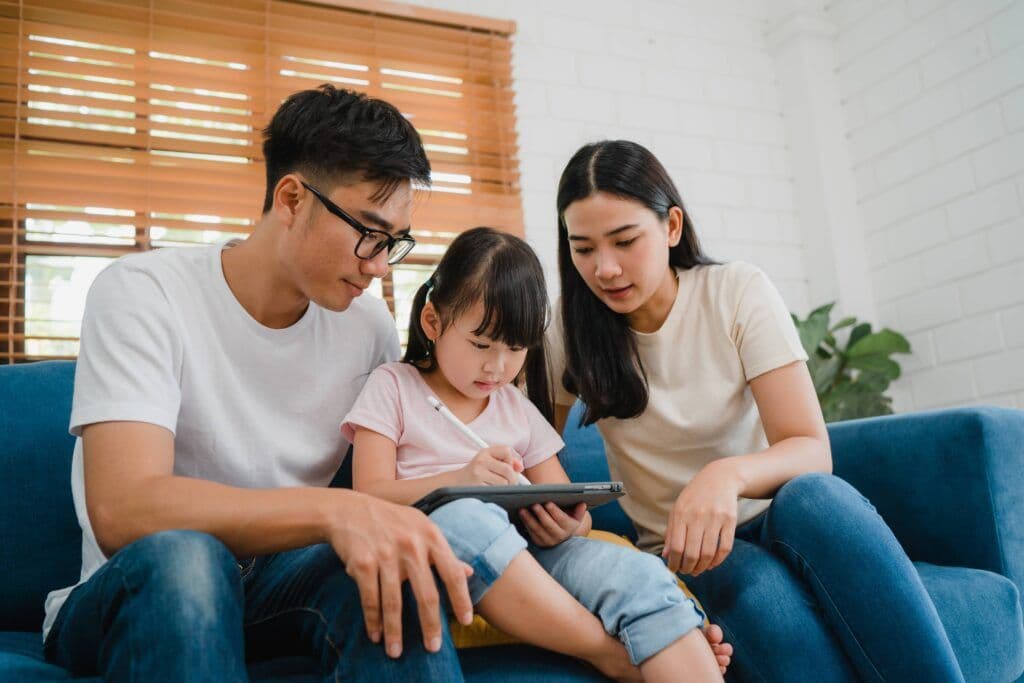 亞裔一家三口坐在藍色沙發上，女兒用觸控筆在平板上畫畫，父母在旁觀看