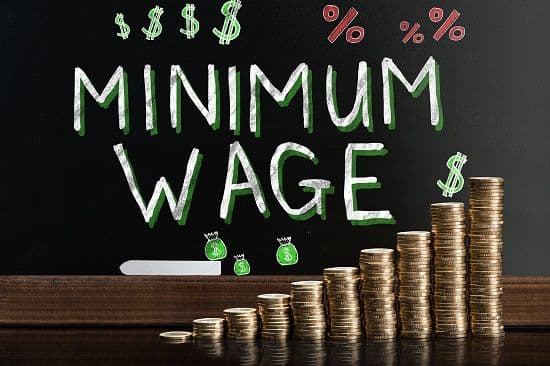 "Minimum wage" chalked on a blackboard behind rising stacks of coins